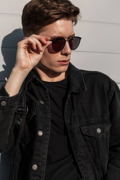 Miejski amerykański facet model mody zakłada klasyczne okulary przeciwsłoneczne na ulicy w słoneczny dzień. Modny portret modny młody człowiek w stylowej dżinsowej czarnej kurtce z fryzurą w pobliżu białego budynku w mieście. - Zdjęcie, obraz