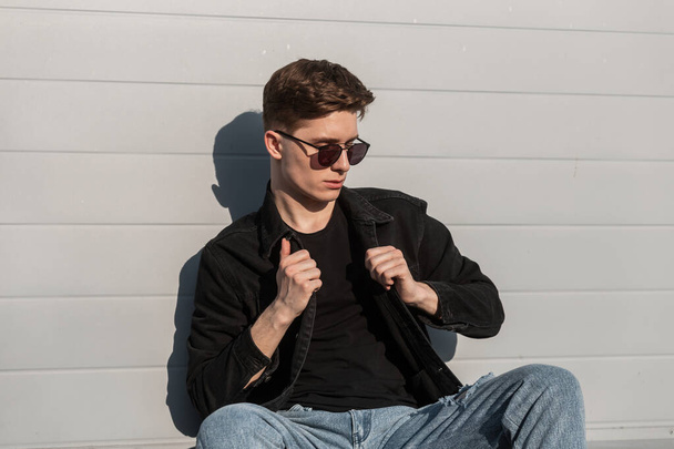 American young stylish hipster man in vintage black sunglasses in fashionable jeans clothes is poses near modern wall in city. Attractive guy fashion model enjoys rarest in sunny day. Casual style. - Foto, immagini