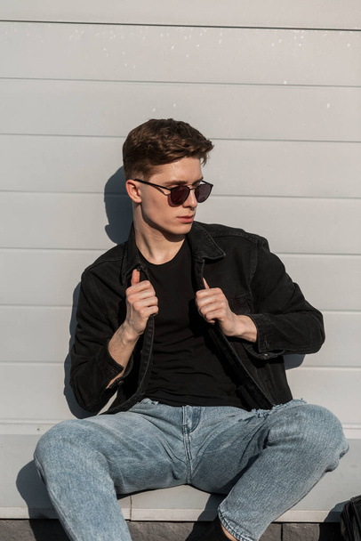 Handsome young man hipster with hairstyle in fashionable sunglasses in blue vintage jeans in black youth denim jacket enjoy sunlight near building in the city. Modern guy on street. Casual style. - Fotografie, Obrázek