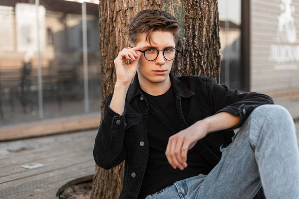 Fashion model young man hipster in stylish jeans clothes straightens trendy glasses in city. American stylish guy in casual denim wear poses near tree on open terrace on street. Youth outfit menswear. - Foto, immagini