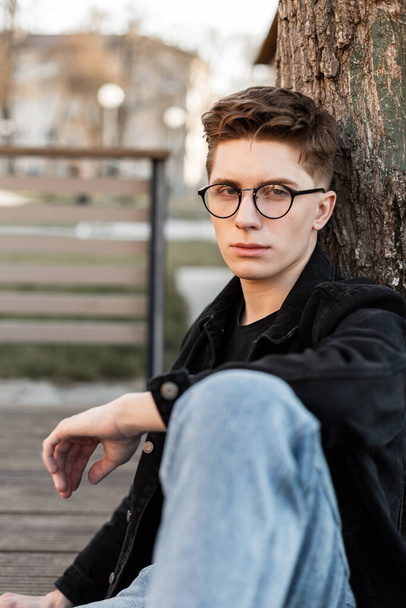 Street portrait american young hipster man with stylish glasses in fashionable denim wear in city. Handsome modern guy in trendy jeans clothes sits near tree outdoors. Casual menswear. Street style. - Foto, Imagen