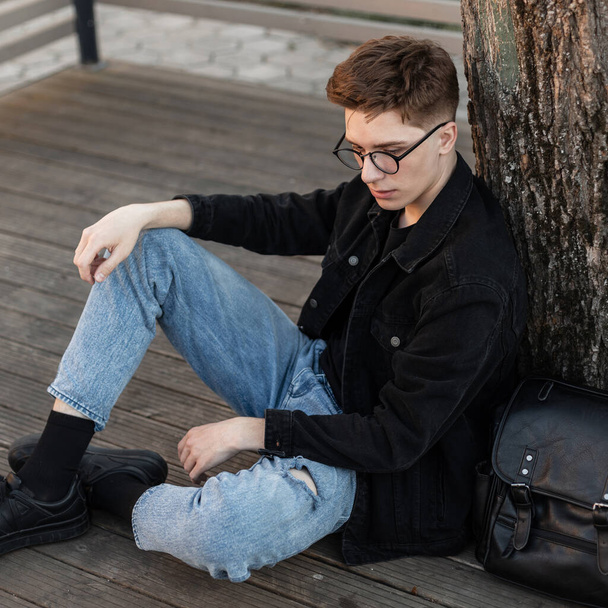 Fashionable young hipster man in vintage glasses in trendy denim casual wear in sneakers with leather backpack is resting near tree on street. Handsome american man in jeans stylish clothes outdoors. - Photo, Image