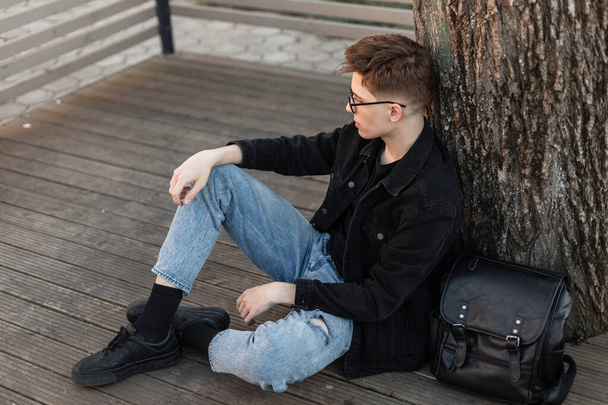 Stylish pretty urban young man in fashionable jeans clothes in fashion black sneakers with leather backpack sits and looks aside near tree outdoors. Trendy guy is resting on wooden terrace in city. - Photo, Image