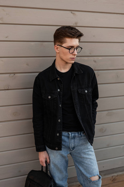 Cool μόδα νεαρός hipster σε γυαλιά με χτένισμα σε κομψό casual denim ρούχα με vintage δερμάτινο μαύρο σακίδιο στέκεται κοντά σε ξύλινο τοίχο στο δρόμο. Αστικός τύπος μοντέλο σε μοντέρνα φθορά σε εξωτερικούς χώρους. - Φωτογραφία, εικόνα