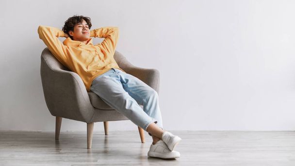 Мирный молодой азиат расслабляется в уютном кресле, ленивый, разбившись о белую стену студии, свободное пространство - Фото, изображение