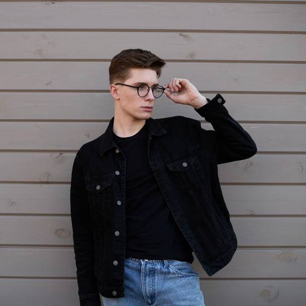 Молодий модний чоловік хіпстер вирівнює стильні окуляри біля дерев'яної стіни на вулиці. Гарний хлопець модель в молодіжному чорному джинсовому піджаку в блакитних джинсах в модній футболці біля будівлі в місті
. - Фото, зображення