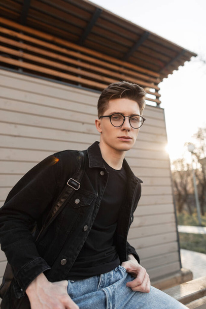 Modern young man hipster in denim black jacket in blue vintage jeans in glasses sits near wooden building on street. Fashionable guy rests on background bright spring sunset. Trendy clothes for men. - Photo, image