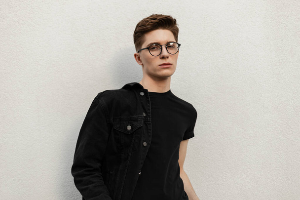 Ein junger europäischer Hipster mit Frisur, modischem T-Shirt, stylischer Jeansjacke und Vintage-Brille ruht sich in der Nähe eines Gebäudes in der Stadt aus. Modelmodel attraktiver Kerl in Jugendkleidung auf der Straße. - Foto, Bild