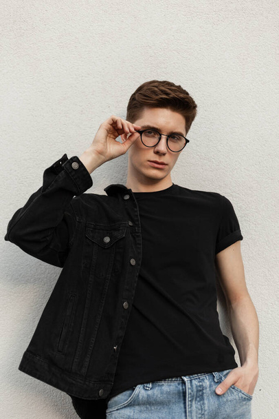 Young fashionable man hipster straightens stylish glasses near vintage wall in street. Handsome guy model in youth black denim jacket in blue jeans in fashion t-shirt near building in city. - Фото, изображение