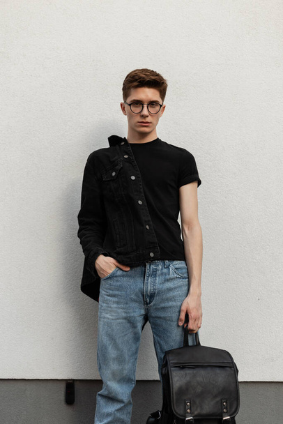 Стильный модный мужчина в джинсовой верхней одежде с рюкзаком у стены - Фото, изображение