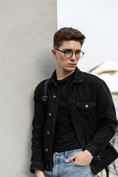 Fashion model serious young man in stylish jeans youth casual wear in fashionable glasses with backpack rest near wall in city. Trendy hipster guy outdoors. New collection menswear. Spring style. - Photo, Image