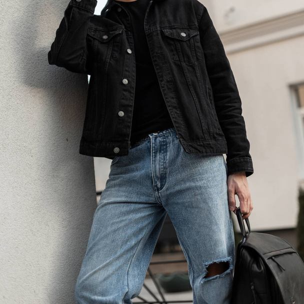 Modieuze knappe man in een zwart denim shirt met een zwart t-shirt en blauwe klassieke jeans houdt een zwarte leren rugzak op straat - Foto, afbeelding