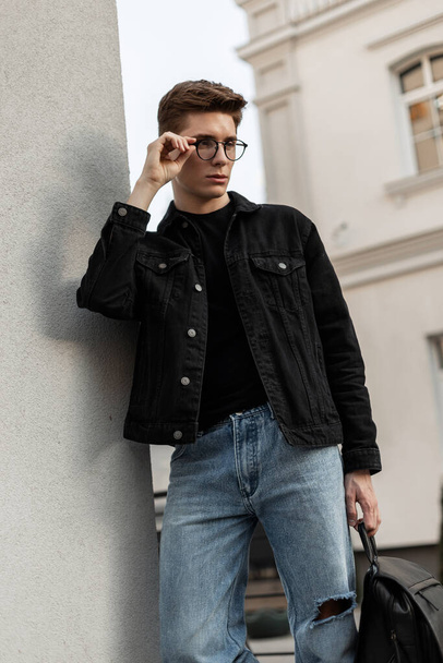 Городской стильный молодой хипстер в джинсовой черной куртке в джинсах с кожаным рюкзаком стоит возле винтажной стены и выпрямляет очки. Модель европейского красавца в модной повседневной одежде. - Фото, изображение