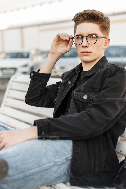 Modern handsome young man in stylish jeans clothes straightens trendy glasses. Urban stylish guy in casual denim wear with trendy hairstyle sits on wooden bench on street. Youth fashion menswear. - Photo, Image