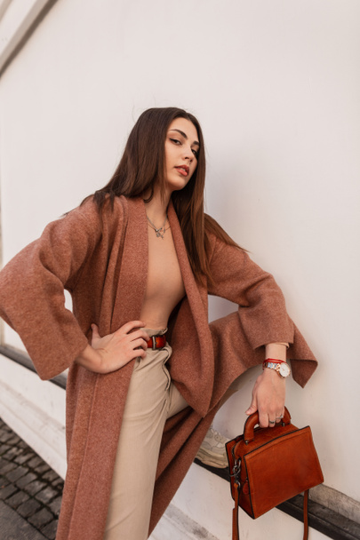 Sexy beautiful fashionable woman in a fashionable coat with a leather handbag posing near the wall in the city - 写真・画像