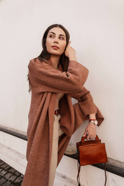 European beautiful young woman in long stylish coat in beige trendy trousers with leather brown fashionable handbag near white vintage building. Elegant girl fashion model posing outdoors in street. - Photo, image
