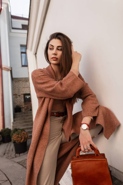 Fashion model elegant young woman in long stylish coat in beige trendy trousers with leather brown fashionable handbag near white vintage building outdoors. Cute urban girl posing outdoors in the city - 写真・画像