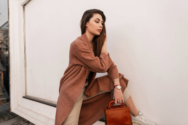 Stylish young woman in fashionable coat in fashion beige pants with brown leather handbag in shoe straightens long hair near vintage wall in city. Pretty elegant girl model posing. Spring style. - Foto, afbeelding