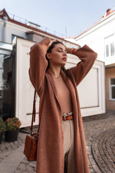 Feminine elegant beautiful young woman in fashionable coat with leather brown handbag poses near white building on street. Sexy girl fashion model straightens chic long hair. Beauty lady outdoors. - Zdjęcie, obraz