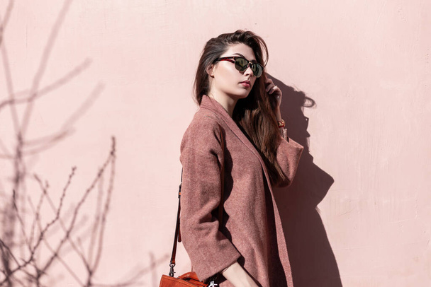 Vintage portrait attractive lovely young woman in dark stylish sunglasses with long hair in elegant coat with handbag near vintage pink wall outdoors. Beautiful gorgeous girl model enjoys spring sun. - Photo, Image