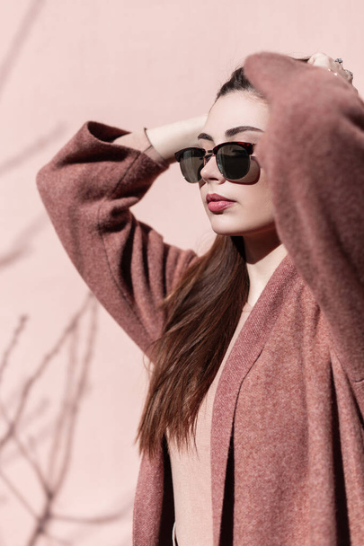 Beautiful portrait young woman in trendy sunglasses in coat outdoors near pink wall on sunny day. Lovely girl with clean skin with sexy lips straightens luxurious hair standing near vintage building. - Фото, зображення