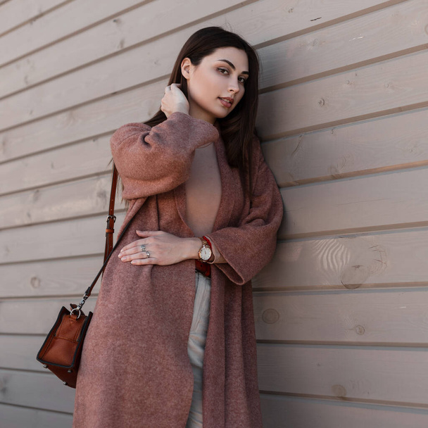 Fashionable young woman model in stylish coat with leather trendy handbag poses near building from boards. Fine cute girl in trendy spring dress is resting near wooden wall on street. Beauty lady. - Photo, Image