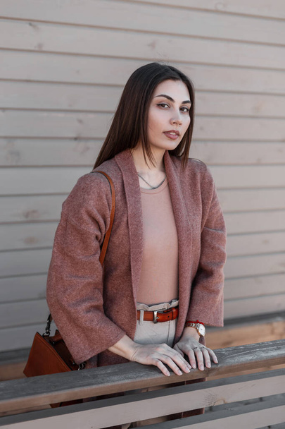 Portrait comely attractive brown-haired young woman in stylish vintage coat with sexy lips near wooden building in city. Fine modern girl in beautiful elegant clothes rests outdoors on spring day. - Photo, Image