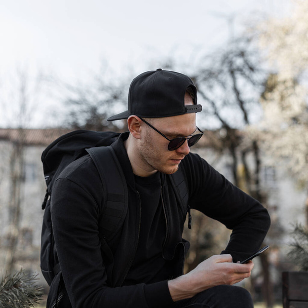 Módní dobře vypadající mladý muž hipster ve stylových slunečních brýlích v čepici v černém oblečení s aktovkou sedí a dívá se na mobilní telefon ve městě za slunečného dne. Atraktivní chlap turista odpočívá na ulici. - Fotografie, Obrázek