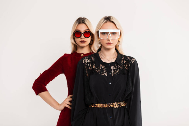 Μοντέρνο πορτρέτο δύο ξανθιές φίλες σε μοντέρνα χρωματιστά γυαλιά νεολαίας σε κόκκινο και μαύρο φορέματα κοντά σε γκρι vintage τοίχο. Σέξι δίδυμες αδερφές με ξανθά μαλλιά ποζάρουν στο δωμάτιο. Κομψά ρούχα για γυναίκες - Φωτογραφία, εικόνα