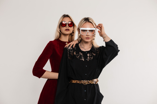 Retrato de moda dos novias adolescentes en gafas de sol juveniles de colores de moda en vestidos rojo-negro cerca de la pared gris vintage. Preciosas hermanas gemelas con el pelo rubio posa en el interior. Elegante atuendo para mujeres. - Foto, Imagen