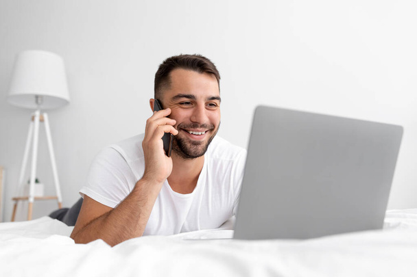 Felice giovane ragazzo attraente con la barba si trova sul letto bianco chiamata per telefono, guarda il computer portatile, di lavoro o di chat - Foto, immagini