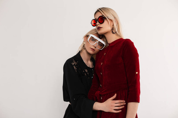 Fresh portrait young attractive blondes sisters women in stylish red-black fashionable dresses in glasses near gray vintage wall in room. Sexy fashion girls posing indoors. Gorgeous ladies hugging. - Photo, image