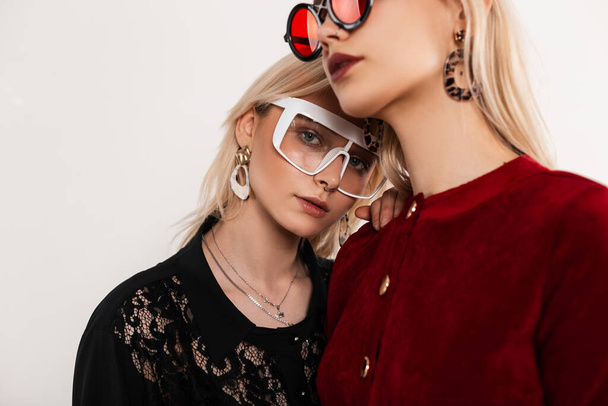 Retro portré fiatal szexi leszbikusok divatos piros-fekete ruhák elegáns szemüvegben állnak egymás mellett szürke háttér a szobában. Divatos lányok elegáns ruhákban pózolnak a stúdióban. Közelkép. - Fotó, kép