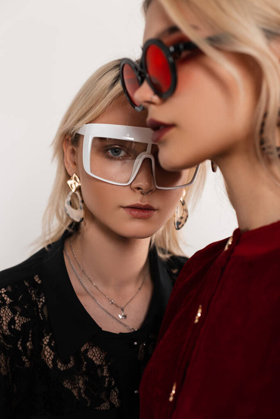 Trendi portré két tinédzser lány, színes szemüvegben, szőke hajjal, piros-fekete ruhában, a szürkeárnyalat mellett állva a stúdióban. Vonzó divatos készletek elegáns viselet pózol belteremben.Közelkép - Fotó, kép