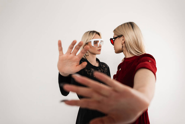 Dwie młode nastolatki w modnych okularach młodzieżowych w stylowych sukienkach zakrywają kamerą dłoń i patrzą na siebie w pobliżu zabytkowej ściany w studio. Atrakcyjne bliźniaki w modnej odzieży pozowanie wewnątrz. - Zdjęcie, obraz