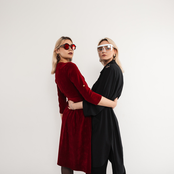 Dwie młode modne dziewczyny dziewczyny z fajnymi okularami przeciwsłonecznymi w stylowych modnych ubraniach pozujących w studio na szarym tle. Dwie piękne nastolatki kobiety z czarnymi i czerwonymi sukienkami wewnątrz - Zdjęcie, obraz