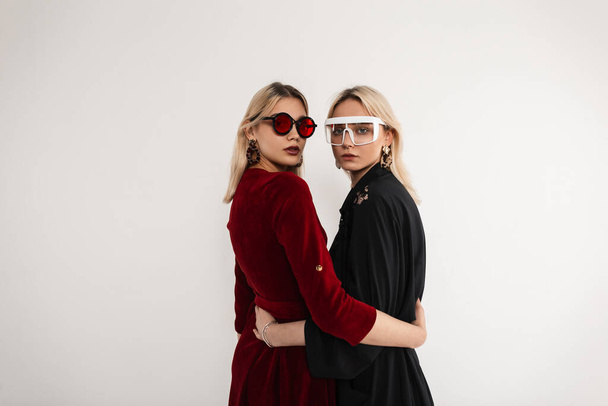 Dwie modne bliźniaczki w okularach przeciwsłonecznych w modnej sukience vintage w pobliżu szarej ściany w studio. Dwie młode kobiety w okularach ubrane w nową kolekcję wewnątrz - Zdjęcie, obraz