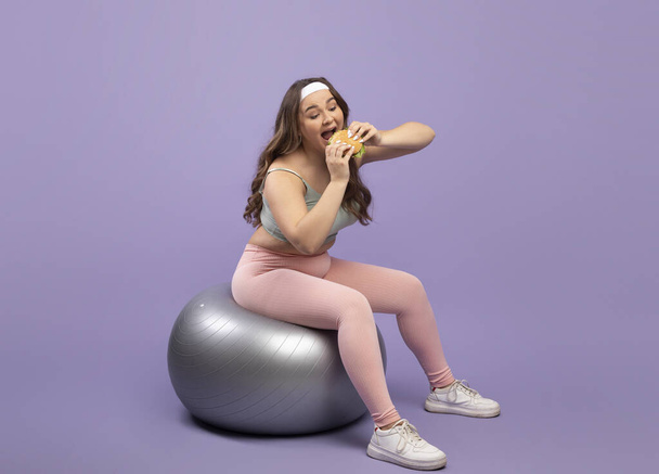 Feliz divertido bastante hambriento caucásico más tamaño joven dama en uniforme deportivo se sienta en la bola en forma y comer hamburguesa - Foto, imagen