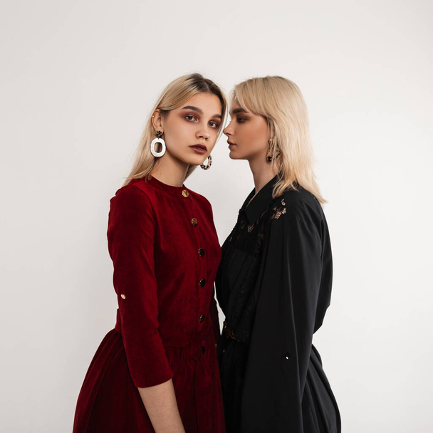 Two young attractive blonde women in fashionable clothes stand in studio near white vintage wall. Lovely girl in elegant black dress looks at beautiful lesbian girlfriend in trendy red dress. Fashion. - Photo, Image