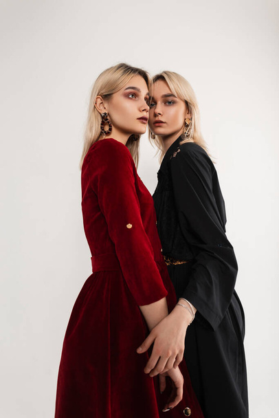 Two young fashion lesbians in stylish black-red clothes posing in studio. Sexy girl fashion model in black dress hugs girlfriend red wear indoors. Youth beautiful clothes in retro style for women. - Photo, Image