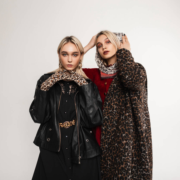 Deux copines européennes branchées dans des vêtements glamour à la mode à l'intérieur. Jolie fille en cuir veste noire avec gants léopard et fille en manteau léopard avec écharpe sur la tête debout près du mur dans la chambre. - Photo, image