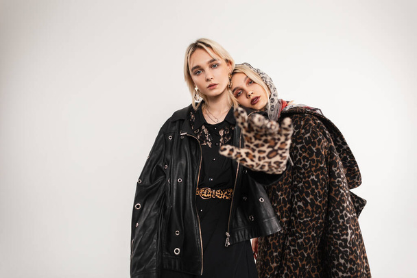 Lovely young attractive girl in leopard glamorous stylish fur coat in scarf hugs woman in leather black jacket in studio near vintage wall. Trendy twin sisters in clothes in retro style in room. - Foto, afbeelding