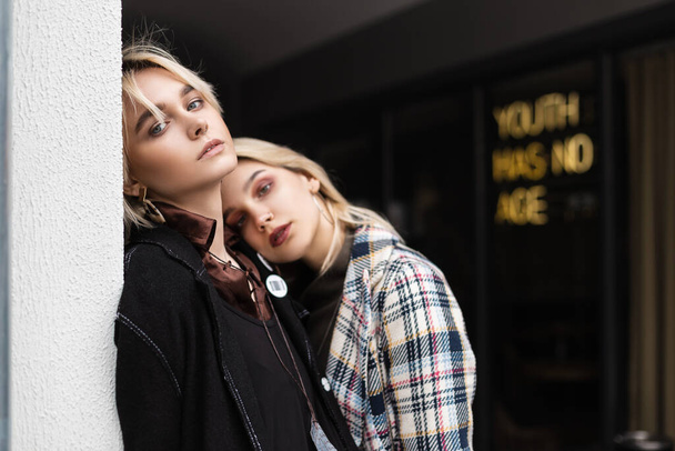 Two young gorgeous blonde women in fashionable clothes stand near vintage wall on street. Beautiful girl in elegant black jacket hugs sexy lesbian girlfriend in trendy checkered blazer. Retro fashion. - Photo, Image