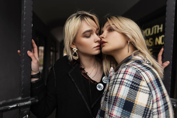 Fiatal gyönyörű nő divatos fekete blézerben megcsókolja barátnő divatos kockás kabát szabadban. Két szexi vonzó leszbikusok elegáns retro csók közelében vas kapu a városban. Divatportré. - Fotó, kép