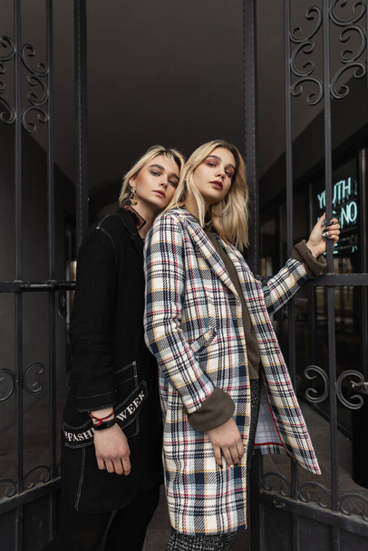 Modern fiatal barátnők divat modell divatos vintage kabátok állnak egymás mellett az utcán. Szexi vonzó lányok gyönyörű kopott pózol közelében vas kapu. Trendi portré két nőről. - Fotó, kép