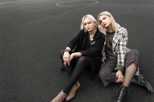 Two european fine sisters twin in trendy casual youth clothes resting on basketball court outdoors. Lovely blonde girlfriends in stylish outfit relax on street in city. Fashion models in trendy wear. - Photo, Image