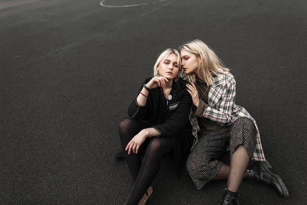 Pretty fashionable young lesbian girlfriends in trendy vintage clothes in retro style sitting next to each other on basketball court outdoors. Cute sexy girls relax on playground. Youth spring wear. - Foto, Bild