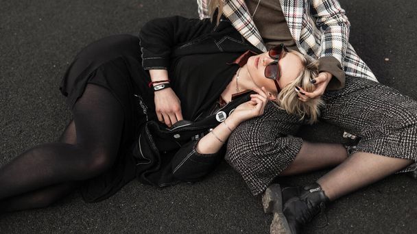 Modern young woman blonde in black stylish clothes resting on asphalt with sister in checkered jacket. Two lovely girlfriends enjoy rest outdoors in city. Casual youth collection women's outfit. - Photo, Image