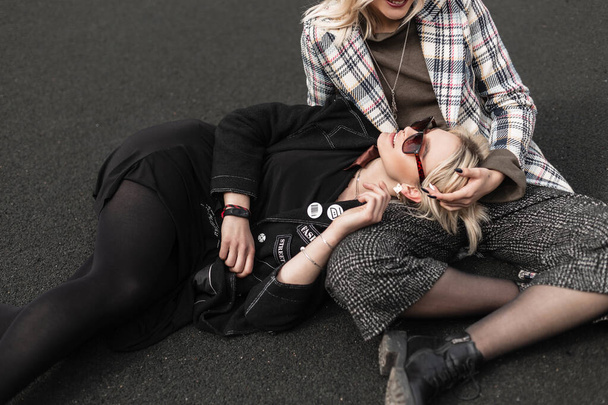 Modieuze jonge vrouw blond in zwarte stijlvolle kleren rusten op asfalt met zus in geruite jas. Twee mooie vriendinnen genieten van de rust buiten in de stad. Casual jeugdcollectie dameskleding. - Foto, afbeelding