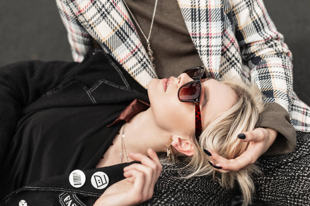 Fresh portrait pretty young blonde woman in fashionable black blazer in stylish sunglasses. Cute girl fashion model in vintage clothes is resting on friend's legs. Trendy sisters relaxes outdoors. - Photo, image